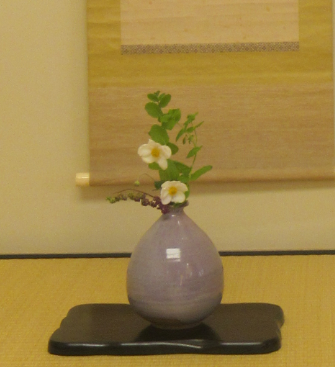 淡い紫色の丸みある花器に白い花と緑の葉が清らかに生けられ、掛け軸を背景に飾られている写真