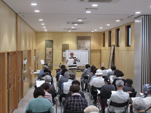 明るい木目調の壁に囲まれた室内で、講師がホワイトボードの前で資料を掲げて解説を行い、それを多くの受講生が椅子に座って熱心に聞き入っている、風流のはじめ館で開催された文化講演会の様子を捉えた写真