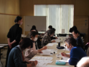 明るい光が差し込む室内で、マスクを着用した多くの参加者が長いテーブルに座り、講師らしき人物が見守るなかで、紙に向かって黙々と墨で文字や絵を描いている写真