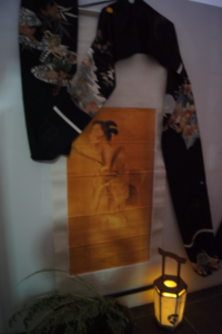 壁に掛けられた黒地の和柄の着物が女性の絵が描かれた掛け軸を囲むように飾られ、足元で灯籠の柔らかな灯りが和の雰囲気を漂わせる写真