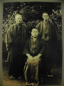 和装の三人が庭木を背に一人を中央に据えて落ち着いた姿勢で整然と並ぶ写真