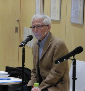 室内で机上の本や飲み物の近くに座った永瀬十悟氏がマイクの前で話す準備をしている様子を捉えた写真