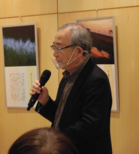 写真展の作品が並ぶ室内で高野ムツオ氏がマイクを手に説明するように立つ様子を捉えた写真