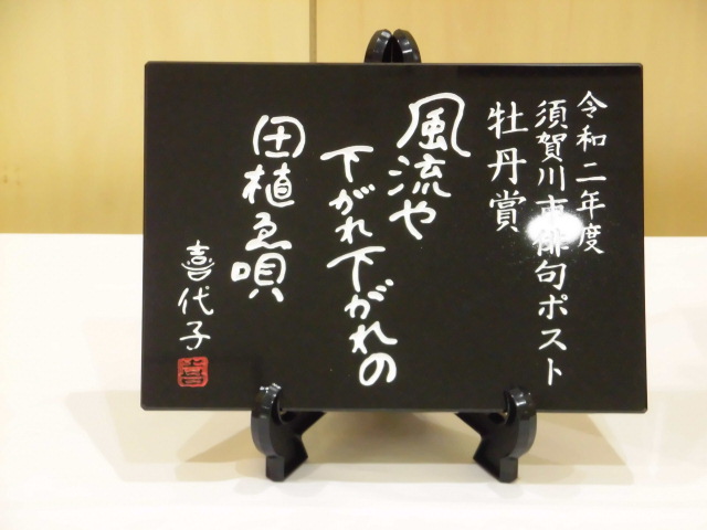 左に「令和二年度 須賀川市俳句ポスト 牡丹賞」と記され、中央には「風流や 下がれ下がれの 田植ゑ唄 喜代子」と白い手書き風の文字で書かれた、黒い光沢のあるプレートがスタンドに立てて展示されている写真