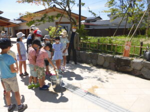 屋外の石垣沿いで、列になって並んだ子どもたちが、バケツから竹の水鉄砲に水を汲んでいる様子の写真