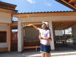 木造の建物の前で、帽子をかぶった女の子が、シャボン玉を飛ばして遊んでいる写真