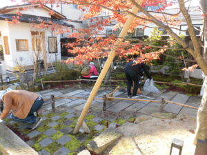 赤く色づいた葉をつけた木がある石畳の庭園で、数人が庭の手入れをしている様子の写真