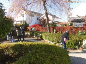 庭園で数人が落ち葉を掃き集めながら植え込みや植栽の間の道の手入れをしている様子の写真