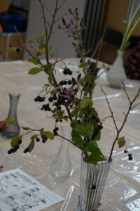 透明シートを敷いたテーブルに置かれた、濃い青の実をつけた枝物や葉を生けた透明の花瓶の写真