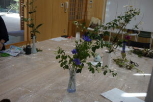 透明シートを敷いた広いテーブルの上に置いた小ぶりの花瓶に緑の葉をつけた枝や青い花が生けられている写真