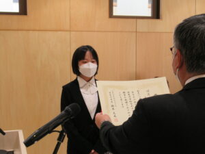 受賞者の女性と向かい合って立ったスーツ姿の男性が、表彰状を読み上げている様子の写真