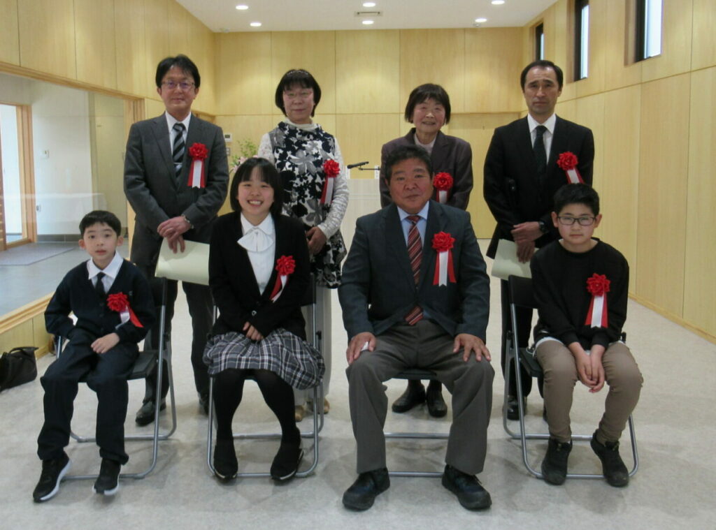 木目調の室内で赤いリボンを胸つけた8人が前列は椅子に座り、後列は立って並んで写した記念写真