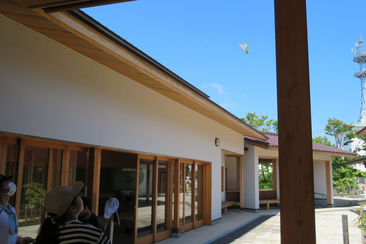 澄み渡る青空の下、和風建築の軒先から、帽子を被った子供たちが空高く舞い上がった紙ひこうきを目で追っている写真