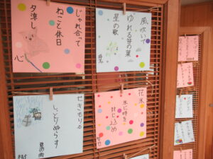 木製のパーティションに、俳句とイラストが描かれた水色やピンク色の四角い紙が整然と貼られ、展示されている写真