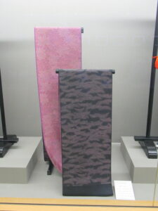 黒い濃淡模様の反物と桃色の反物が台座にかけられて前後に並んだ展示の写真