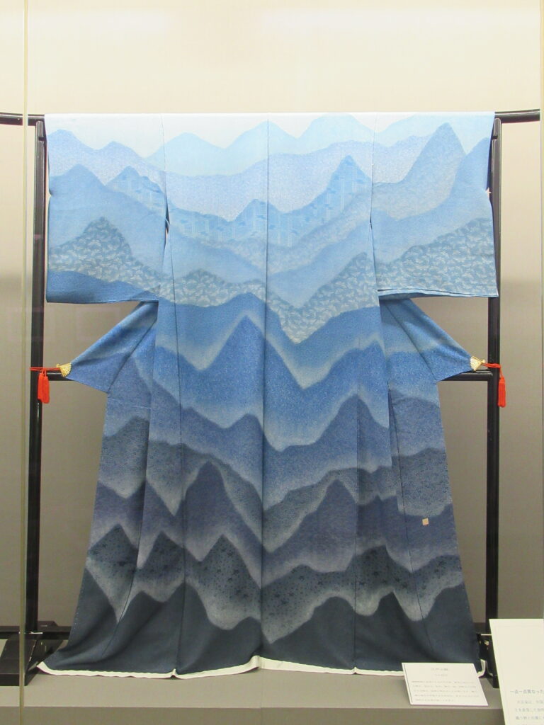 衣桁に掛けられて展示された、青の濃淡で重なる山並みの文様が広がる着物の写真