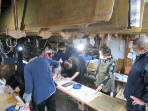 天井に複数の薄い板が下げられ、複数の作業台が並んだ工房で、たくさんの参加者が染色の体験をしているワークショップの様子の写真