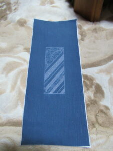 青色の生地の中央に斜線と花のような模様を組み合わせた長方形がデザインされた、長方形の敷物の写真