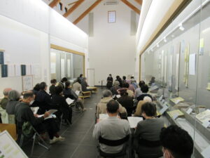 天井の高い白い壁の展示室で、多くの来場者が資料を手に着席し、前方の講演者の話を聞いている写真