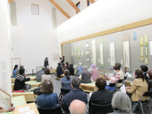 右側の壁に掛け軸などが展示された室内で、ベンチやいすに座った参加者たちが、前方の講演者のトークに傾聴している様子の写真