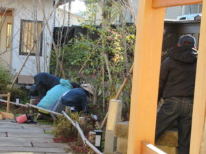 石畳横の木の柵の中で、複数の人が植栽の手入れをしている後ろ姿の写真