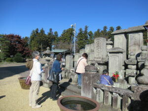 たくさんの古い石碑が並ぶ中、3つ並んだ大きな石碑の前に並んだ数名の男女が、花などを供えてお参りをしている様子の写真