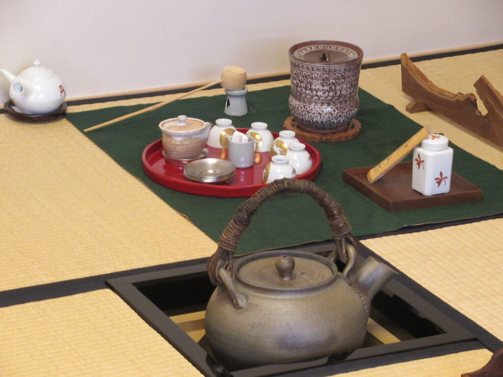 畳の上に敷かれた緑色の敷物の上に、湯飲みなどが乗った赤い盆や柄杓などが並び、手前の炉には茶釜が置かれている様子の写真