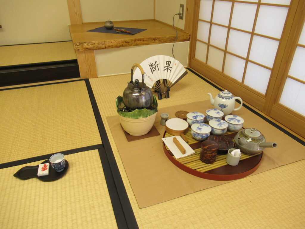 畳の上に飾られた扇子の前に茶釜と急須や湯呑みが乗った丸盆が置かれ、右側には湯飲みと菓子が乗ったひょうたん型のお盆が置かれている写真