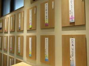 室内の壁に整然と並んだ木製パネルに、俳句の短冊が掛けられて展示されている写真