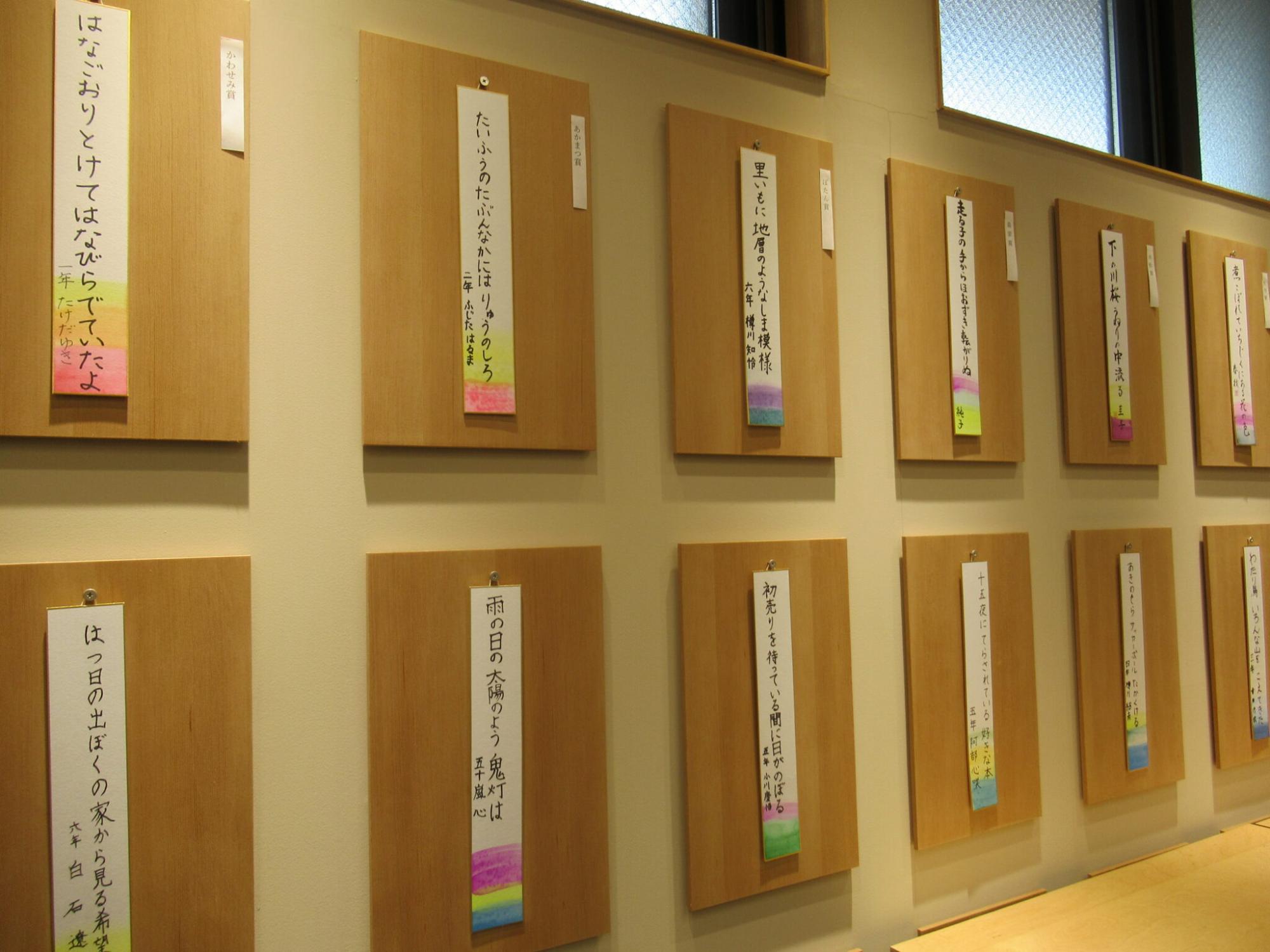 壁に掲示された多数の木製の板に、色鮮やかな装飾が施された俳句の短冊が掛けられて展示されている写真