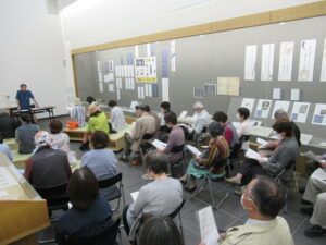 展示室に並んだ椅子に、たくさんの参加者が資料を手に座り、前方に講師の男性が立っている会場全体を後方から撮影した写真