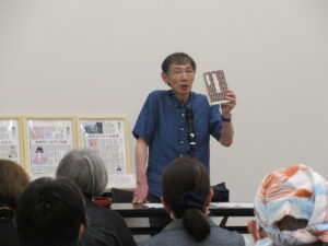 参加者の前に立った講師の男性が、片手に冊子を掲げながら話をしている写真