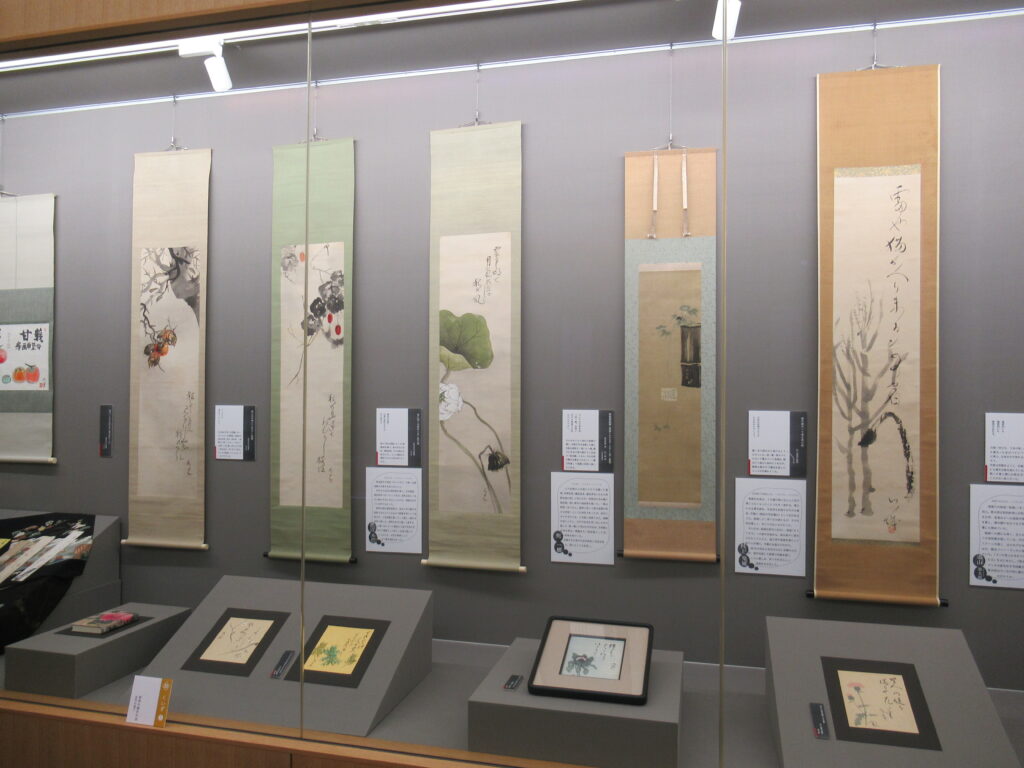 花鳥画に俳句を添えた、掛軸や色紙の俳画作品がガラスケース内に展示されている写真