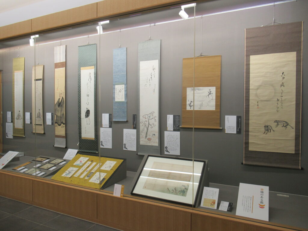 複数の掛軸や短冊などの俳画作品がガラスケースに展示されている展示風景の写真