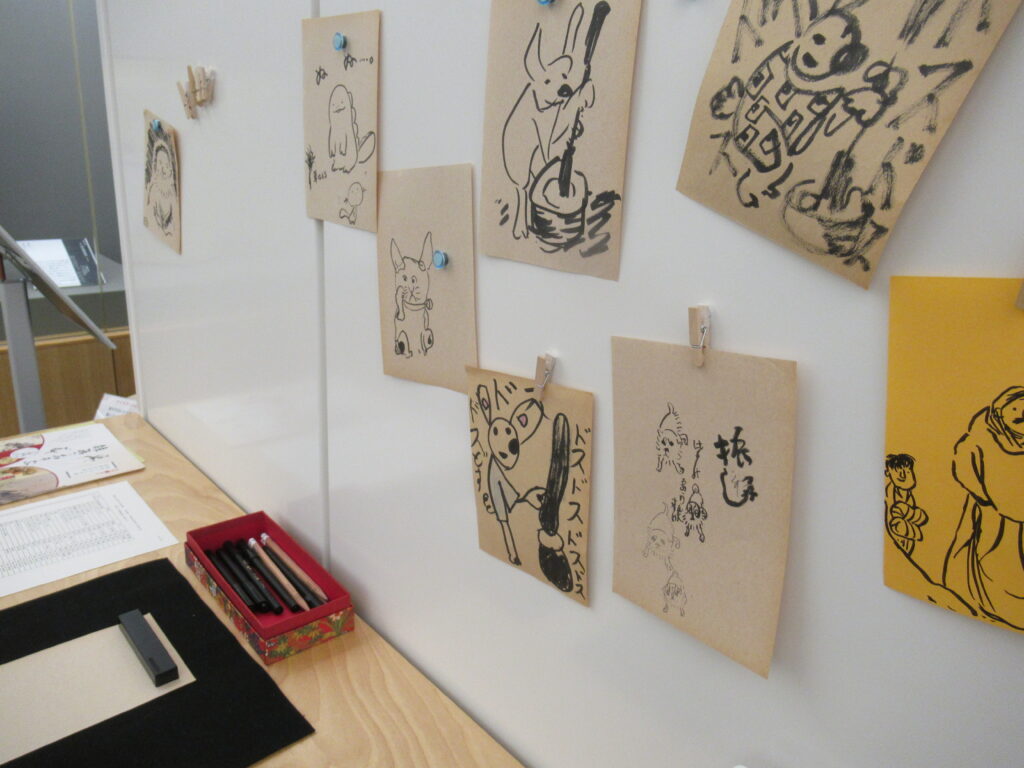 白い壁に、餅をつくウサギなどの絵が描かれた小さな紙が複数飾られ、その下の台には筆記具が並んでいる写真