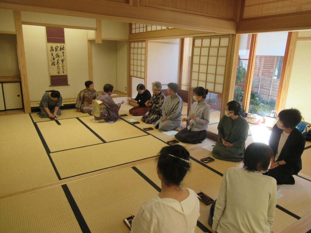床の間に掛け軸が掛けられた明るい和室で、正座で並んだ参加者たちと向かい合って座った和装の女性が、作法を教えている様子の写真