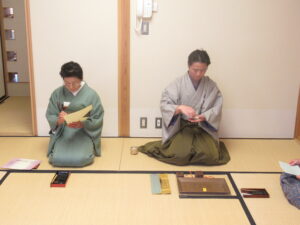 畳の上に置いた筆記用具や香の道具を前に、和装の男女が正座している写真