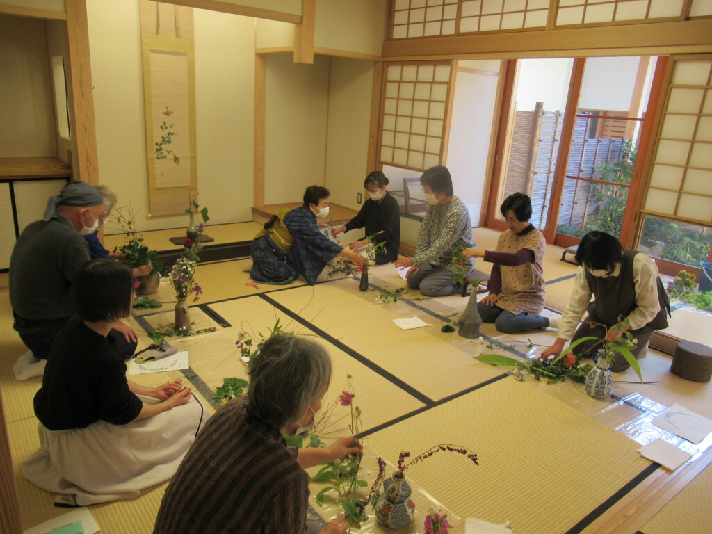 和室で向かい合って座った8人の参加者たちが畳の上に置いた花瓶に草花を生け、講師の女性が一人ずつ指導をしている様子の写真