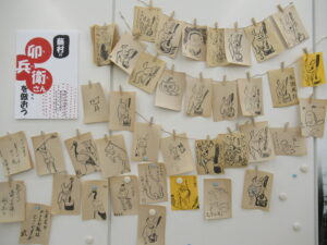 「卯兵衛さんを倣おう」のチラシが貼られた壁に、絵が描かれた複数のカードがところせましとクリップで留められている写真