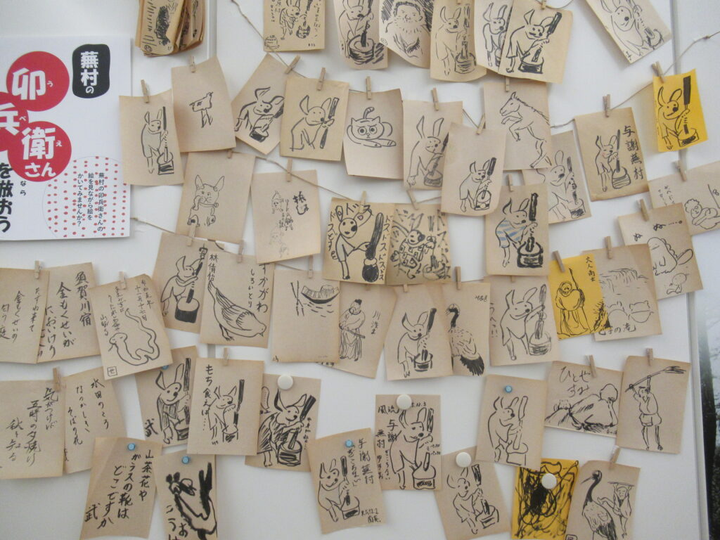 「卯兵衛さんを倣おう」のチラシが貼られた壁に、餅をつくウサギや人物などが黒で描かれた、たくさんの正方形の小さな紙が、重なりながら壁一面にクリップやマグネットで留められている写真