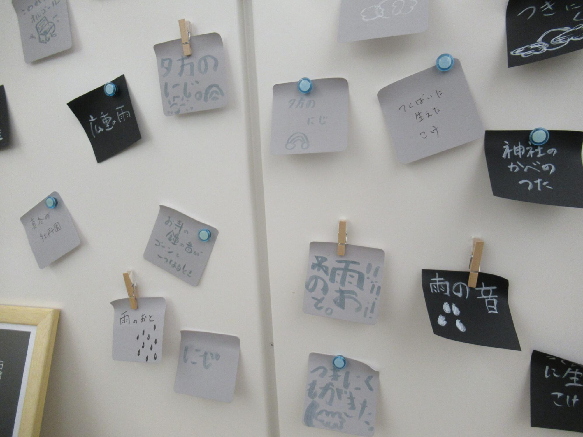 白い壁に、雨や虹などの言葉や絵が描かれた、複数の黒とグレーの付箋紙がクリップやマグネットで貼られている写真