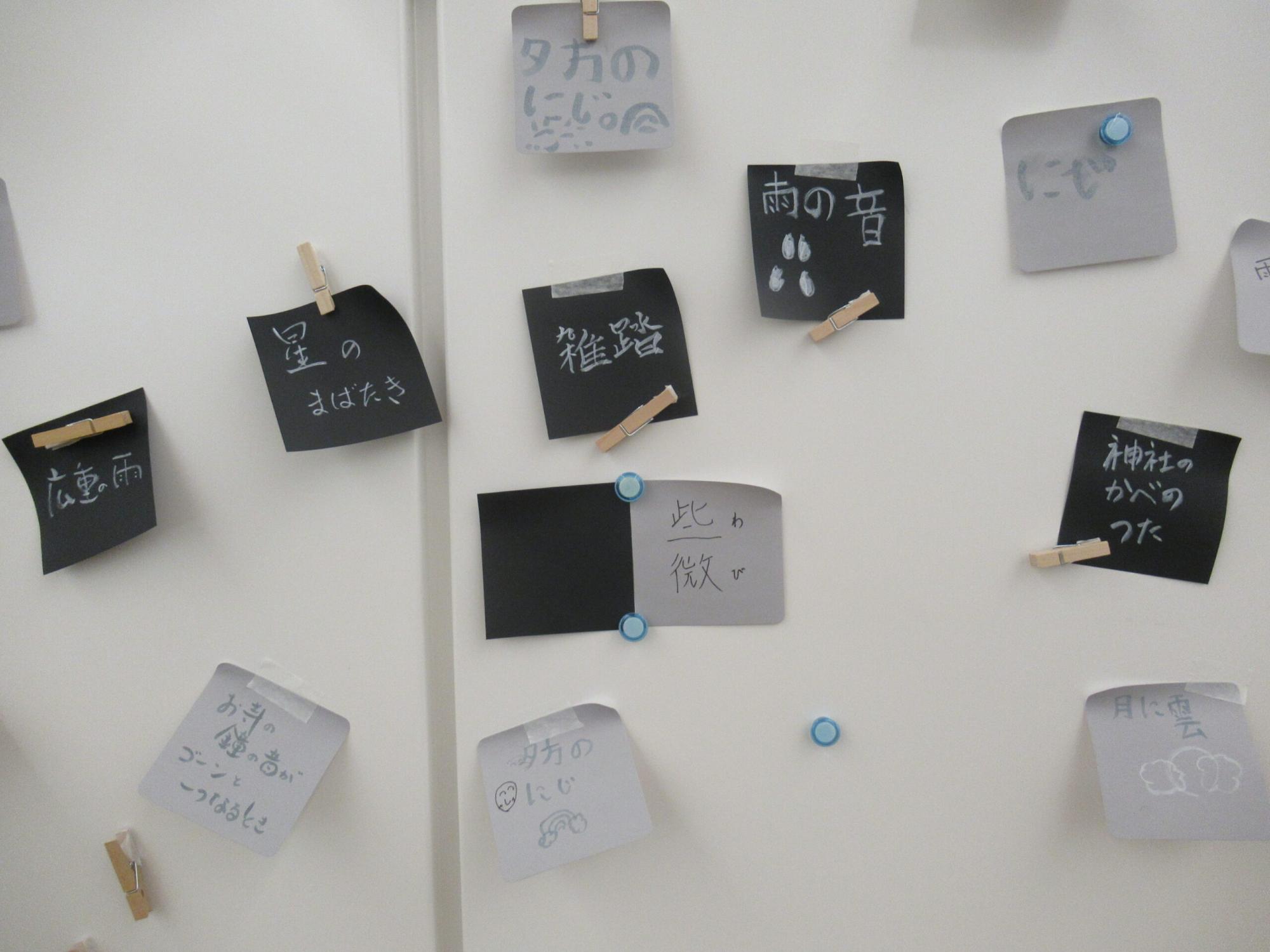 白い壁に雨や星、虹などの言葉や絵が描かれた黒とグレーの付箋紙がクリップやマグネット、テープで貼られている写真