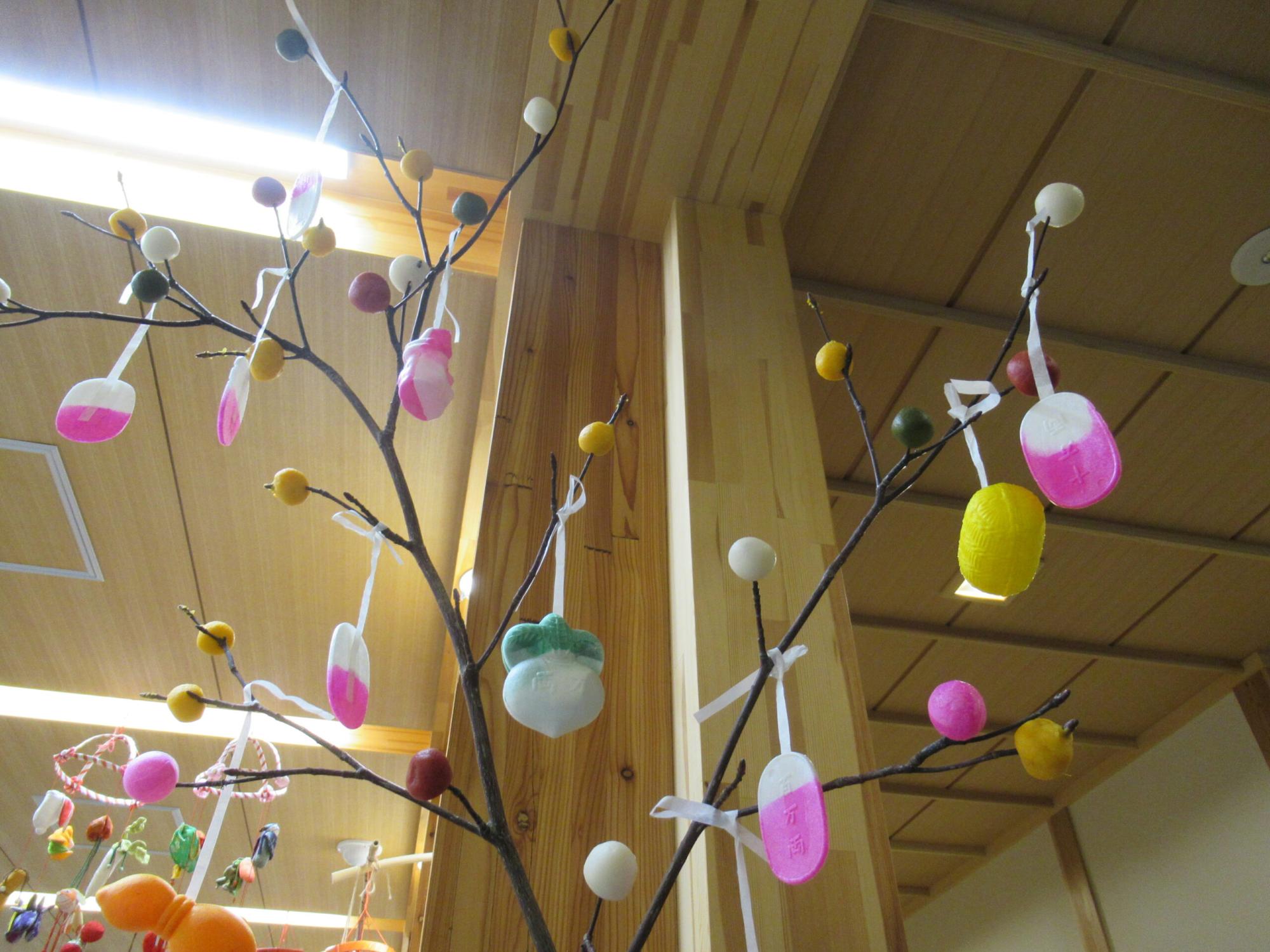 木の枝に白や桃色、黄色などのだんごが刺され、カブなどの形をした飾りが吊るされただんごさしを下から見上げて撮影した写真