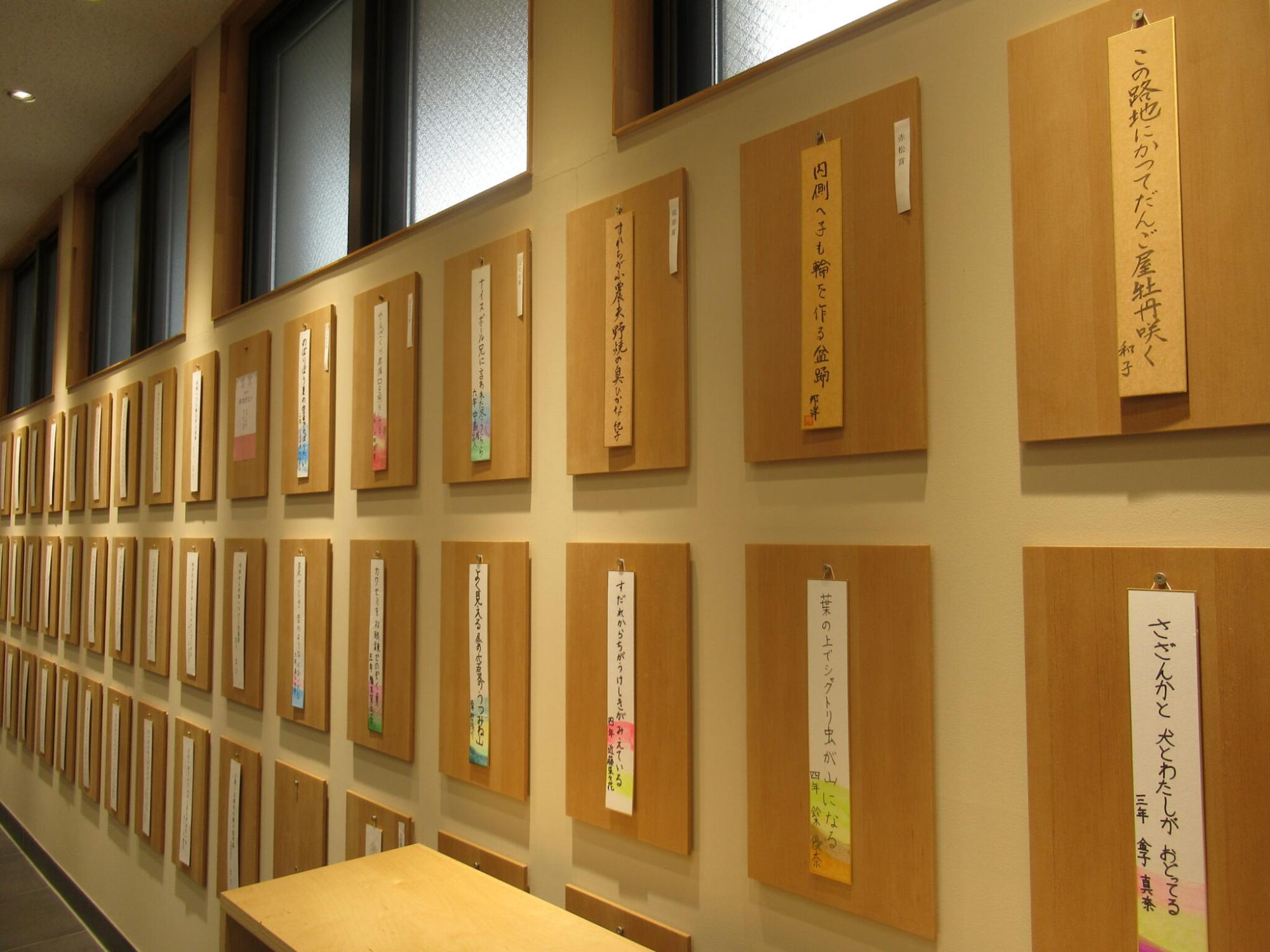 明るいベージュの壁一面に、手書きで俳句が書かれた短冊が掛かったたくさんの木製のパネルが規則正しく何列にもわたって展示されている様子の写真
