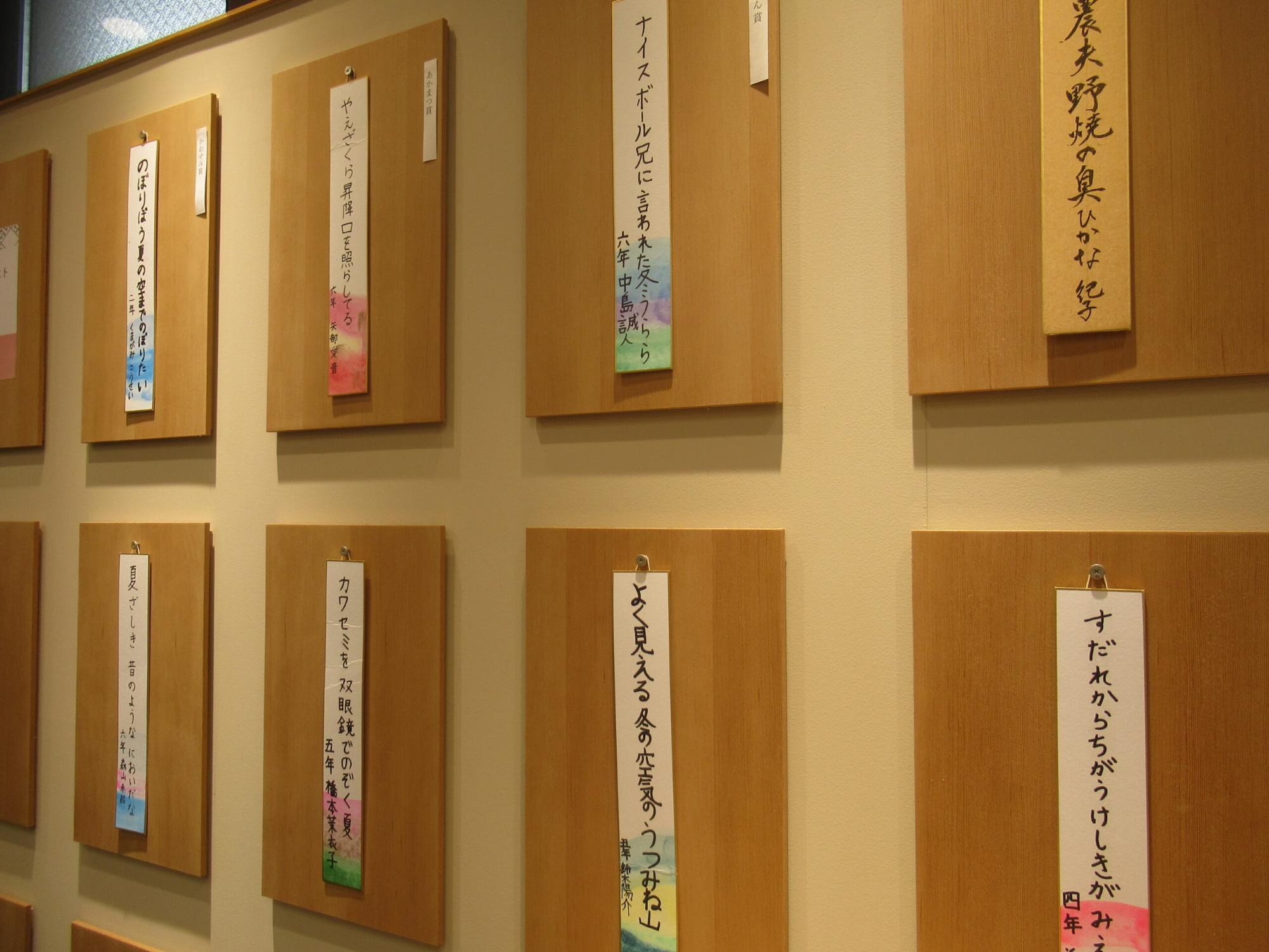 俳句が手書きされた短冊が掛けられた木製パネルが、壁に整然と並んで展示されている写真