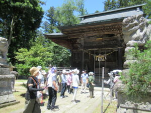 木々に囲まれた古い社殿前に立ち並び説明を聞いている参加者たちの写真