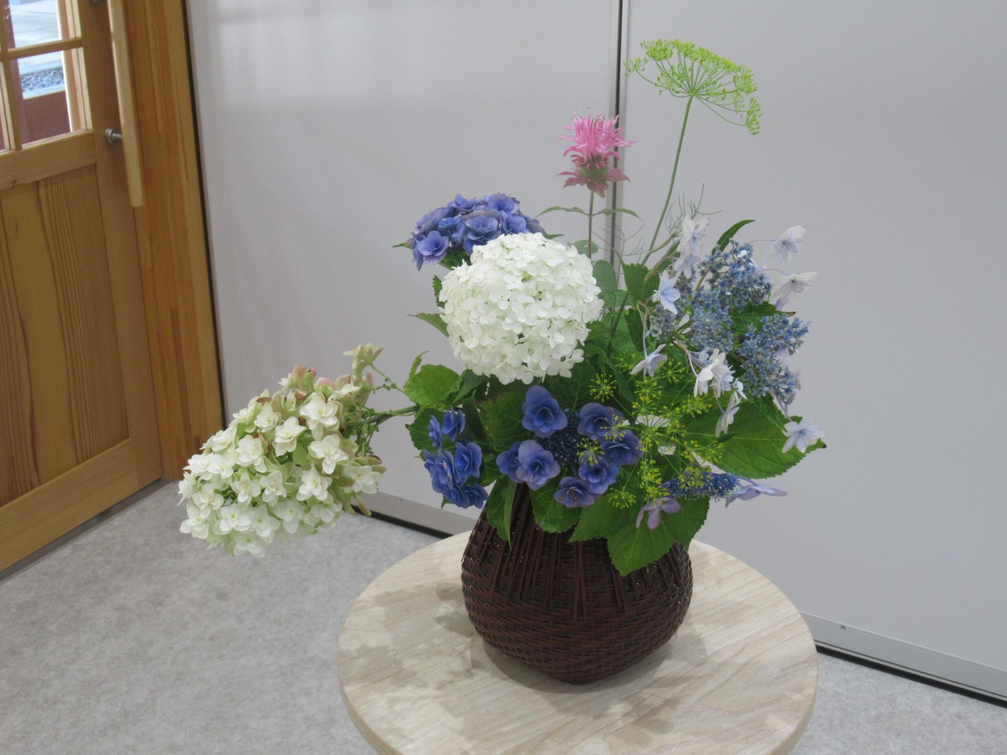 茶色の編み籠のような花瓶に、白や青、紫色の紫陽花を中心に、ピンク色の花や繊細な黄緑色の植物がバランスよく活けられ、丸い木製の台の上に置かれている、室内を彩る華やかな生け花の写真
