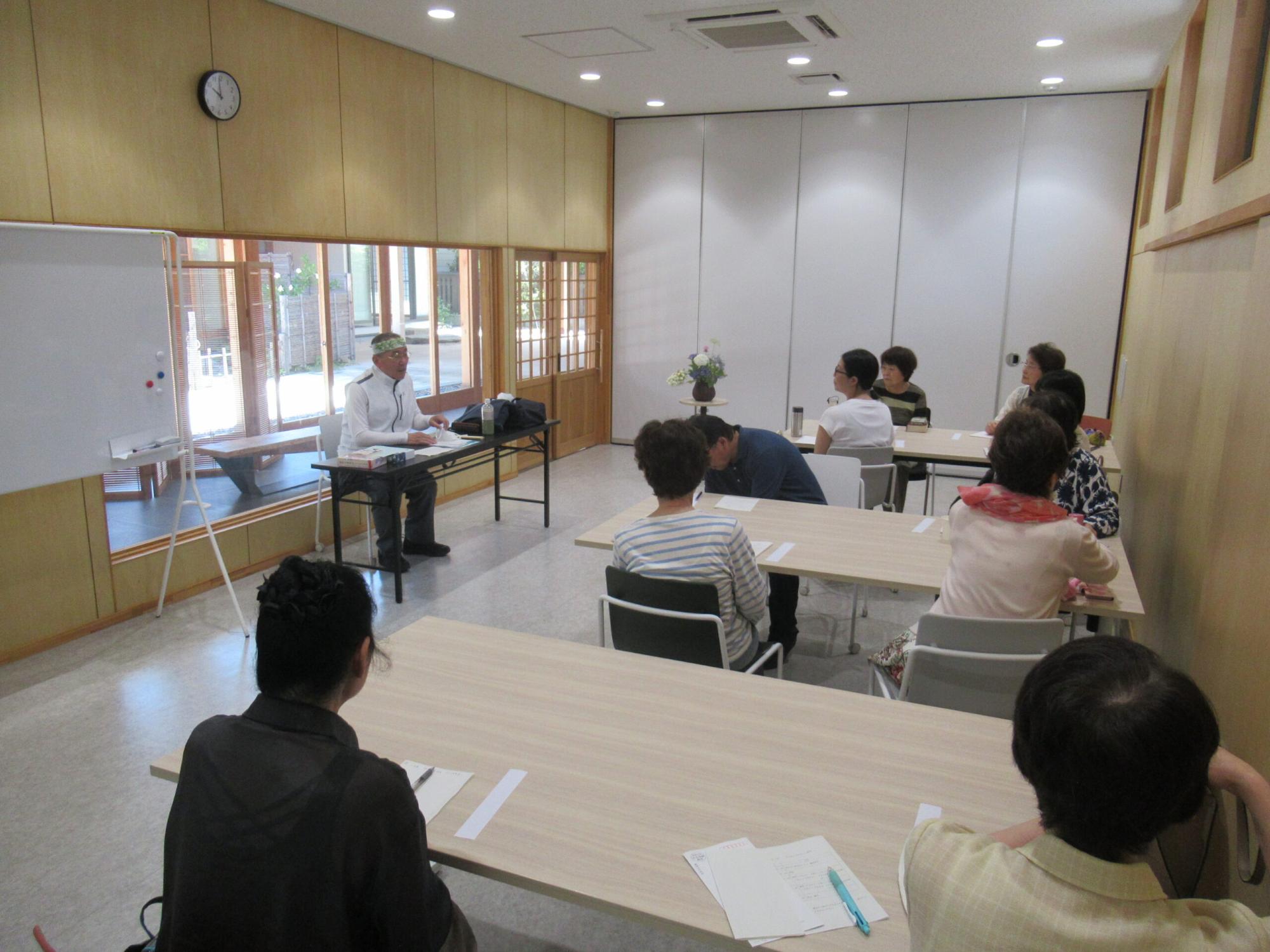 明るい会議室のような室内で、前方の長机に座った講師の男性が、複数名の受講生が座る机の方を向きながら、手元の資料に基づいて熱心に講座を進めている様子を後方から捉えた写真