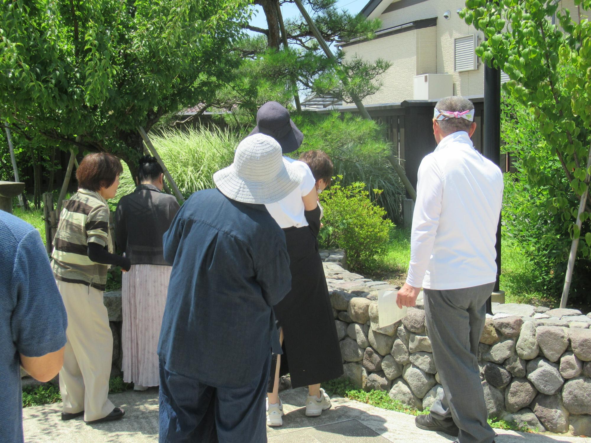日差しが降り注ぐ庭園で、帽子を被った女性たちと白いシャツ姿の男性が、石垣の向こうにある植栽を熱心に眺めながら散策を楽しんでいる様子を捉えた写真