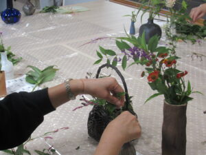 細長い花瓶に赤や紫の花が生けられ、その横に置いた黒い籠型の花器に、緑の葉や紫の花を丁寧に挿して整えている女性の手元を捉えた写真